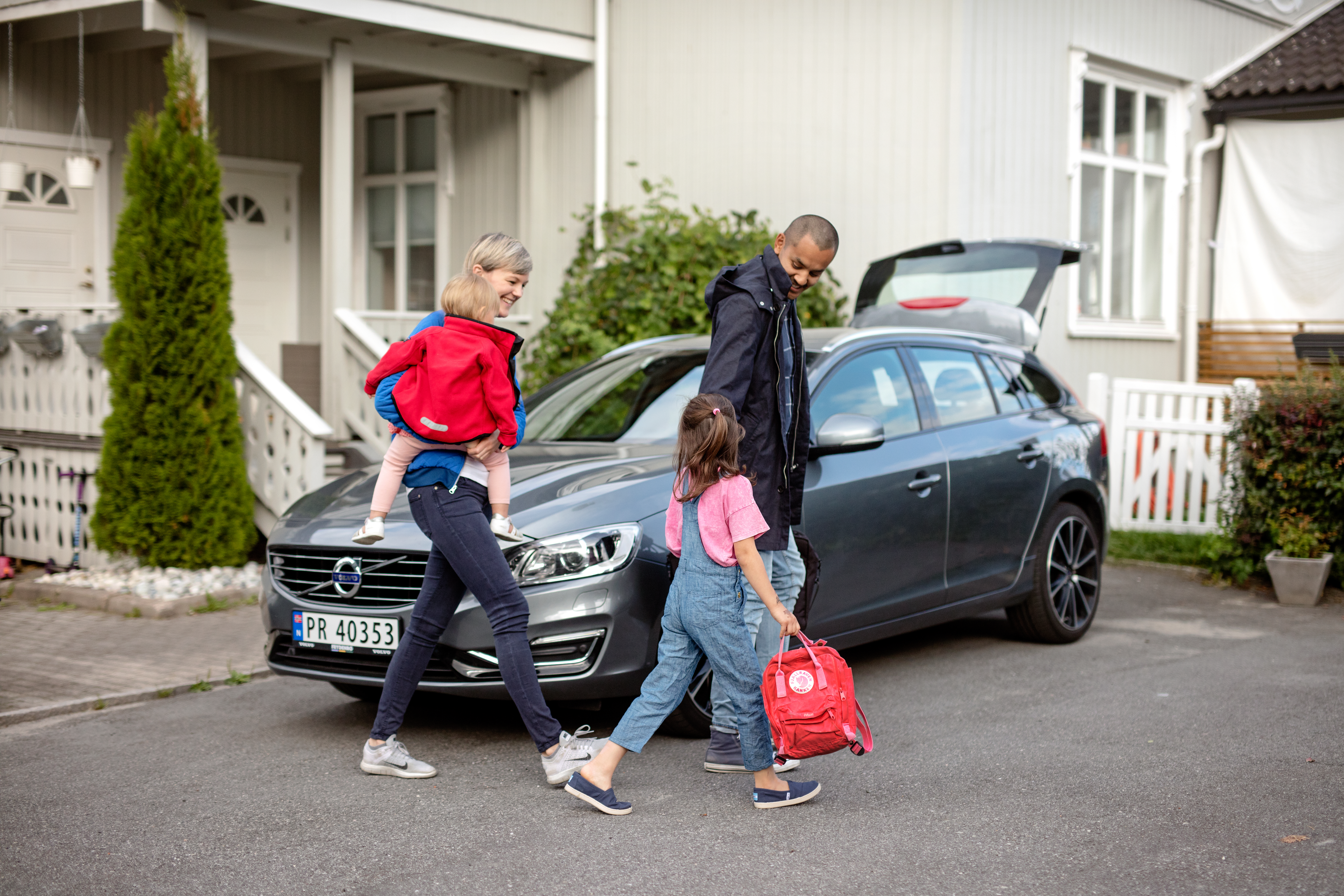 Barnefamilie på vei til å sette seg inn i familiebilen som står parkert utenfor stort hvit hus.