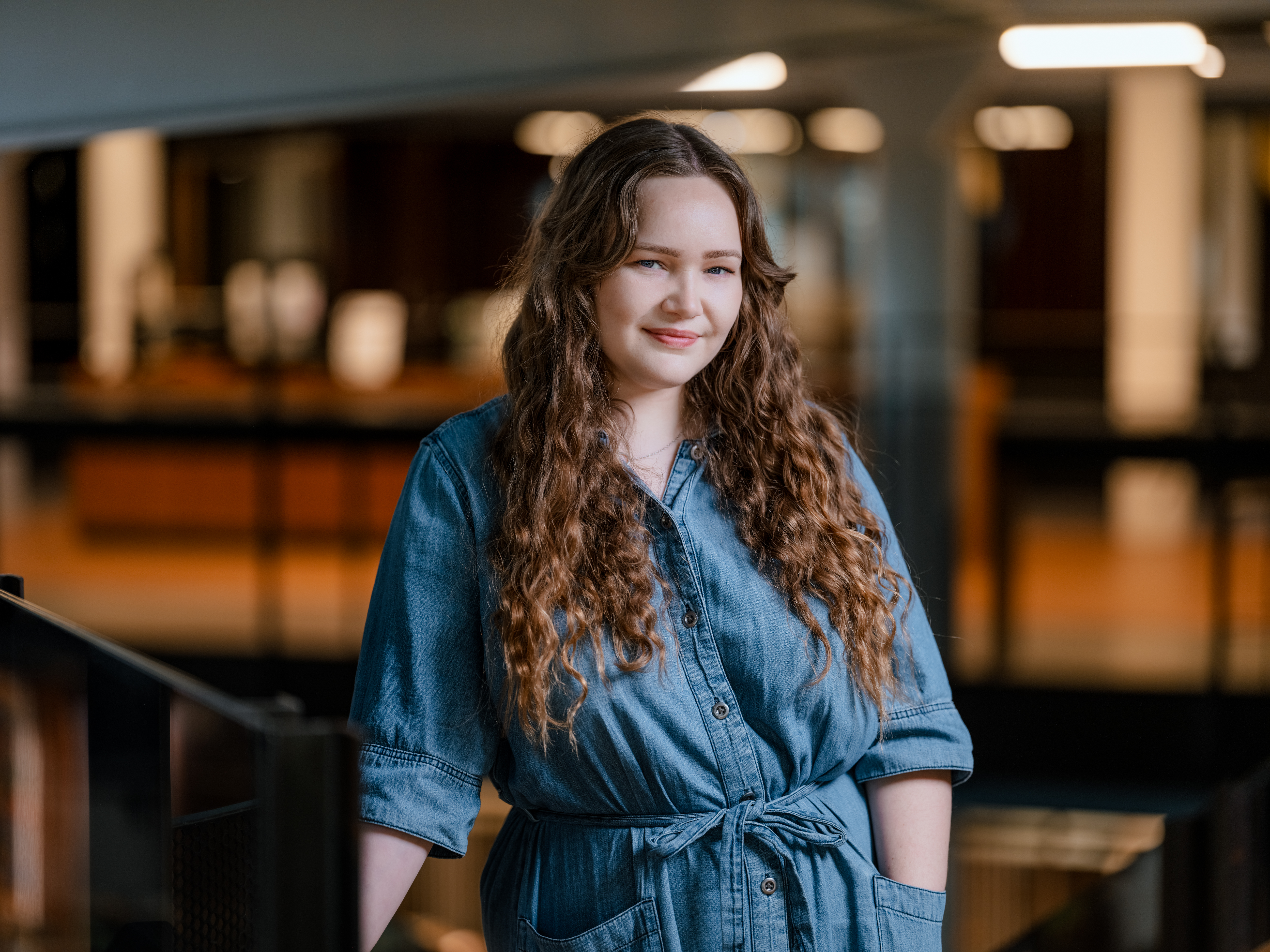 Ung kvinne ser mot kamera. Lener seg på rekkverk inne i et bygg. Foto.