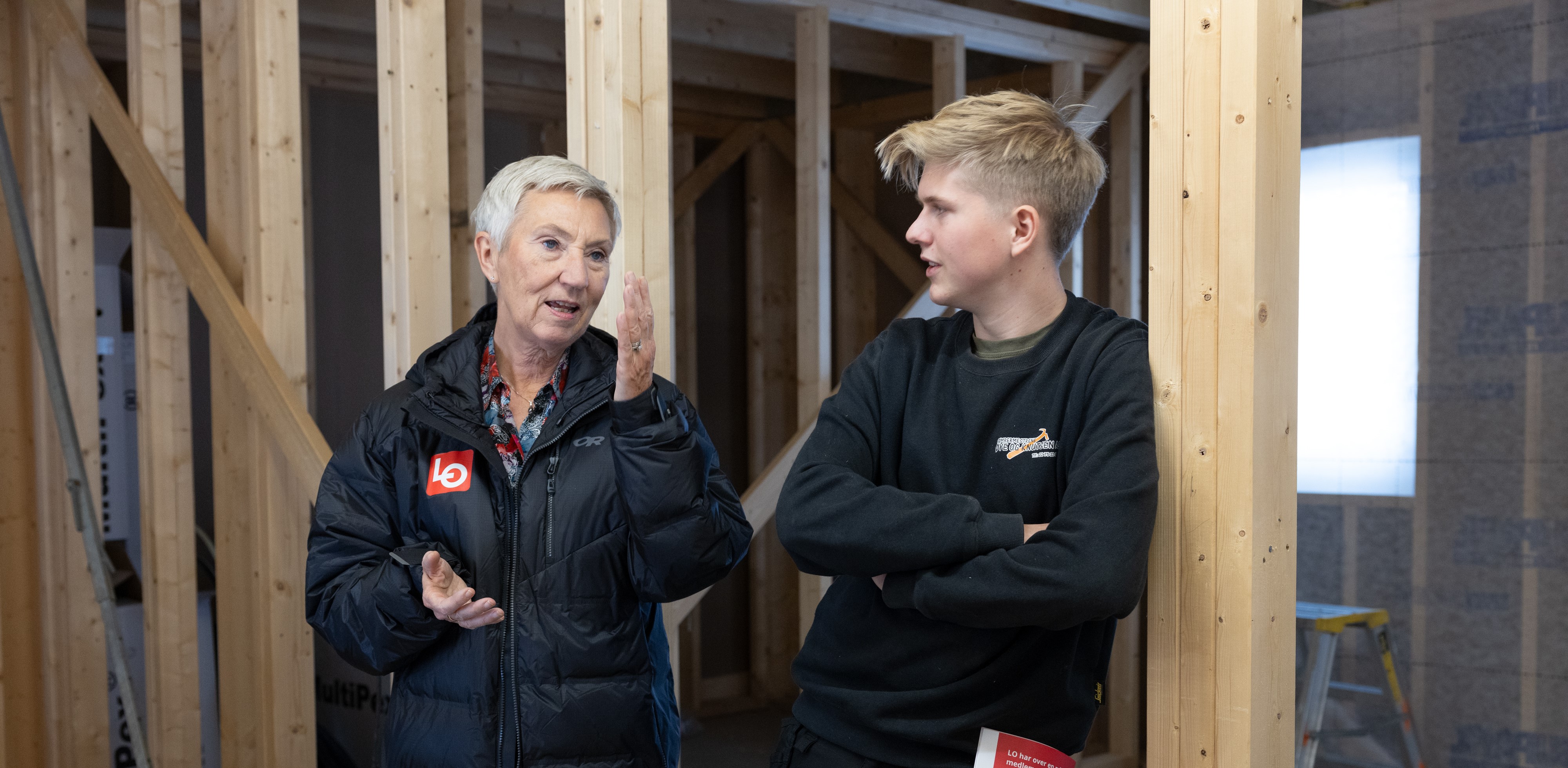 Peggy Hessen følsvik i enusiastisk dialog med en ung lærling på en byggearbeidsplass.