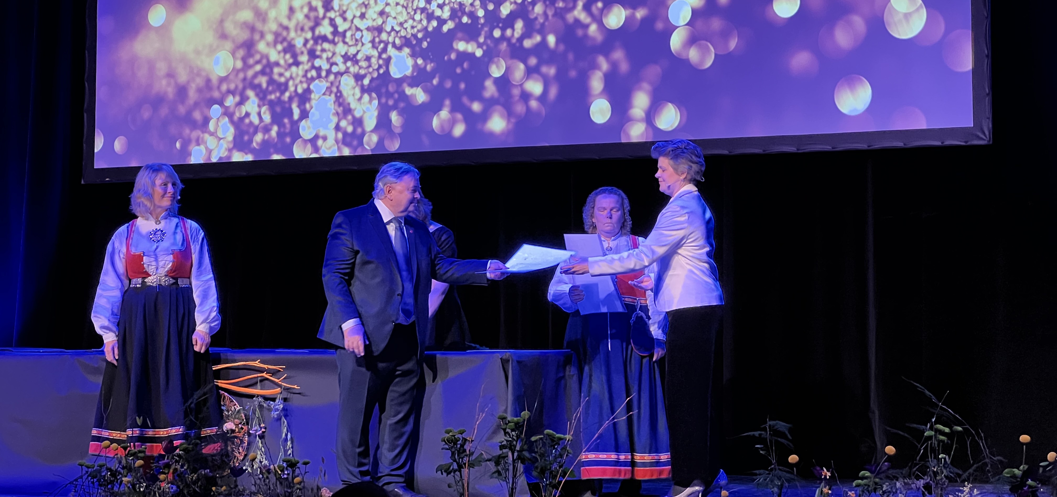 Mennesker på en scene, ikledd bunad - klare til å dele ut fagbrev