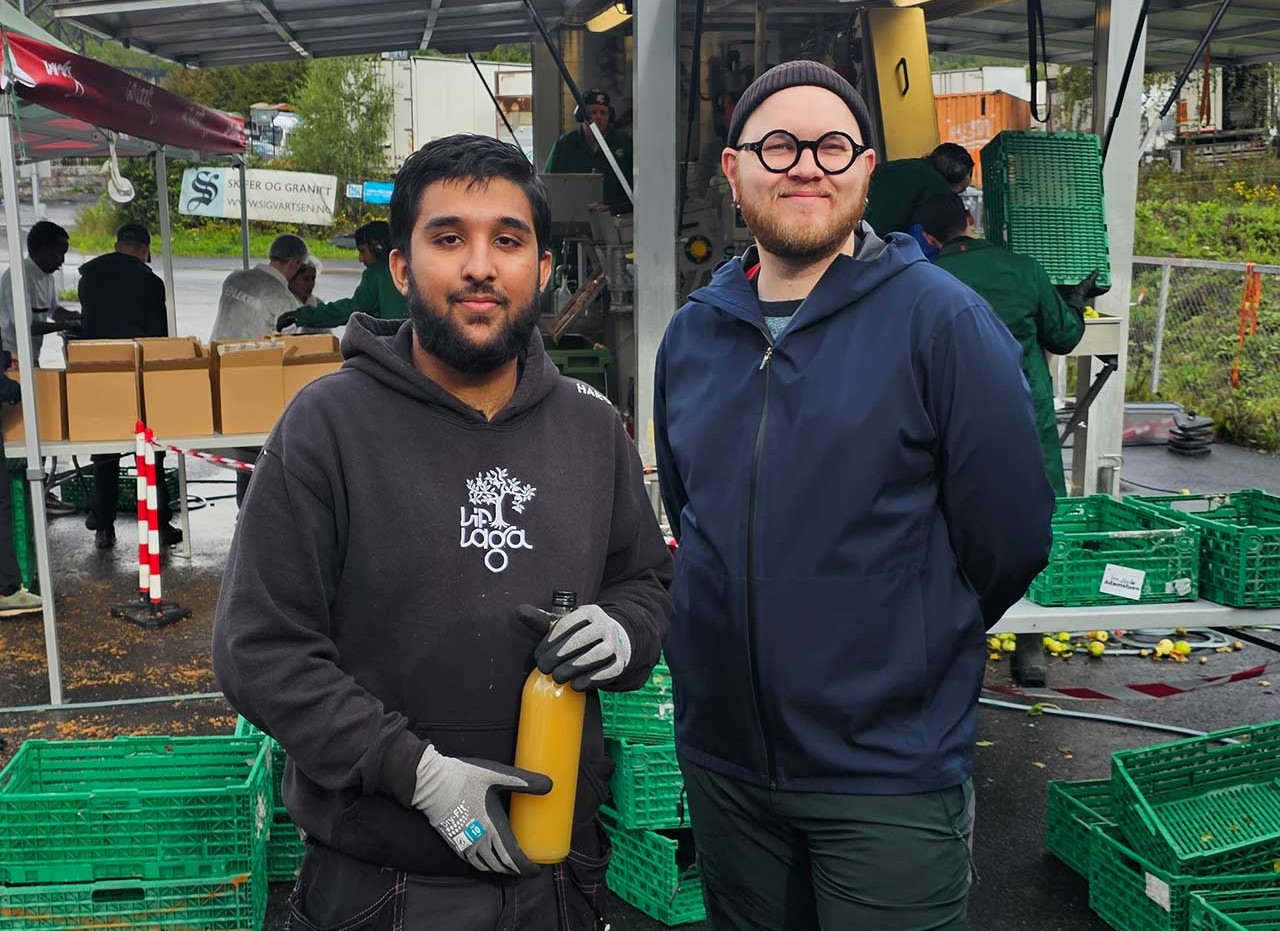 Ung mørk gutt står på vesntre side og holder en glassflaske med eplemost. Kristoffer Jamessen, lys mann med briller, står til høyre