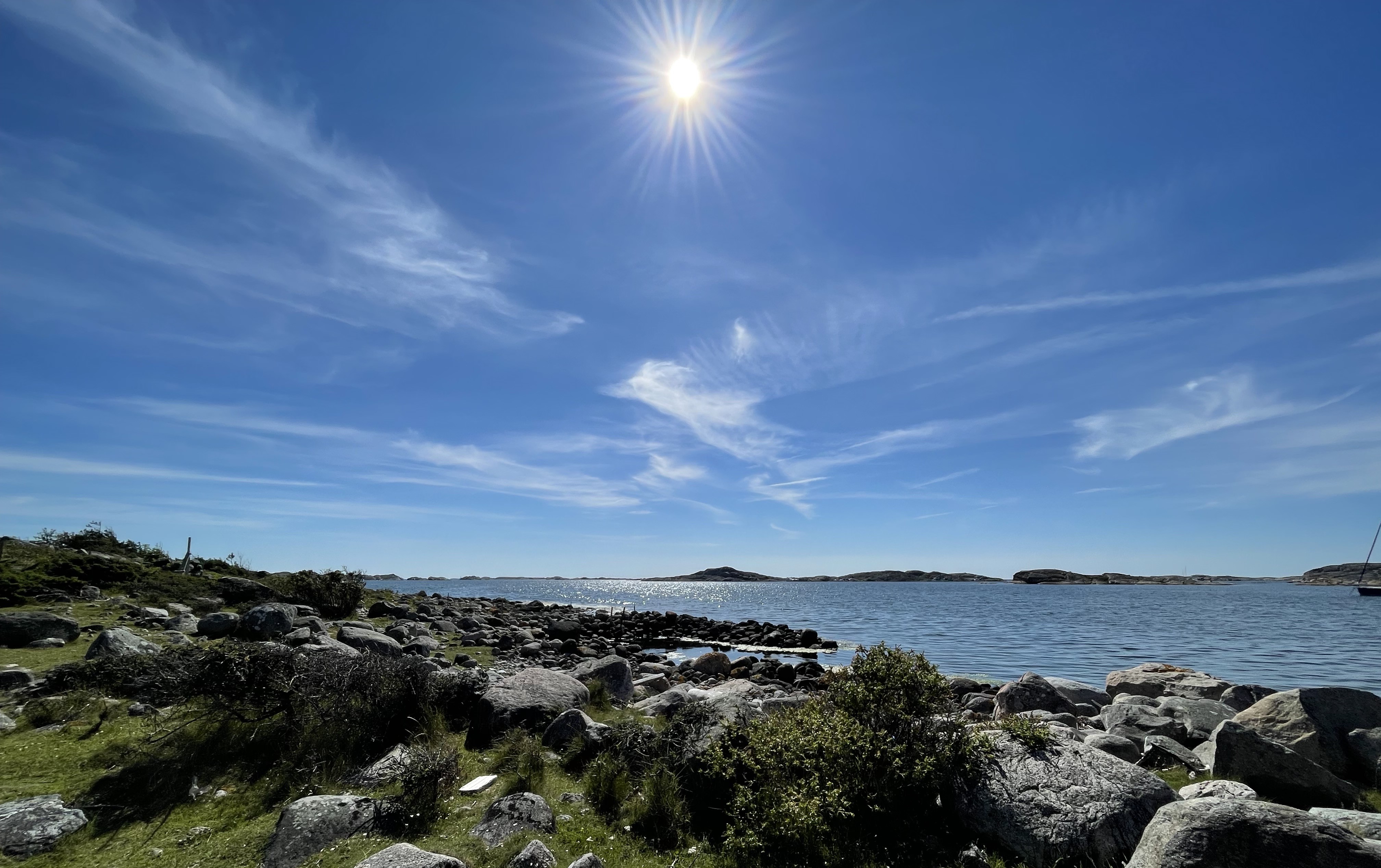 Man ser ut over havet med blå himmel med noen få lette skyer og en sterk sol. Stener som går ned i vannet.