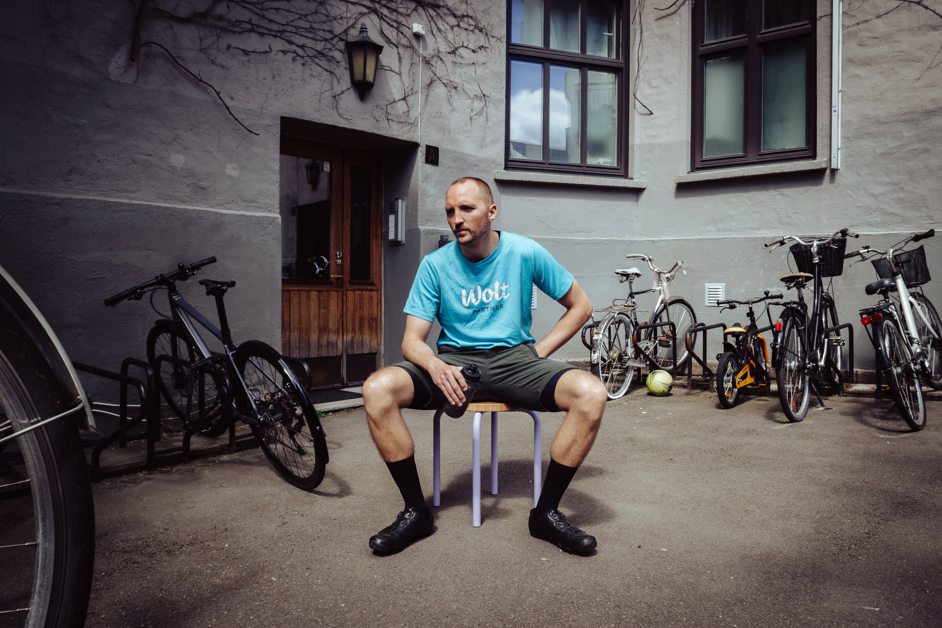 Sykkebud Espen Landgraff sitter på en stol i en bybakgård med sykler rundt seg iført blå Wolt-tskjorte. Foto