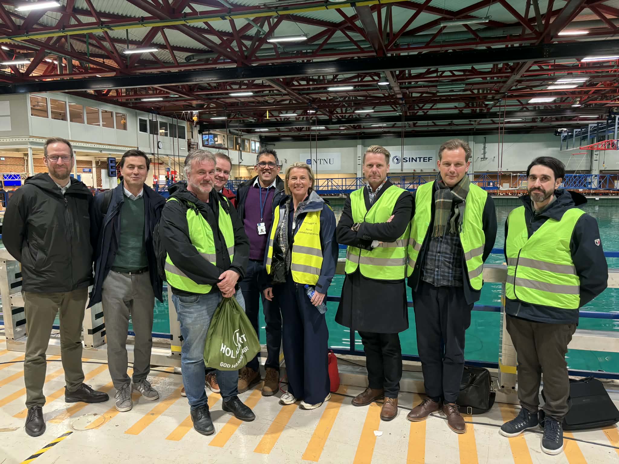 Delegasjon fra LO, NTNU og Sintef poserer i gule vester og vernehjelm sammen i industrilokale. Foto.