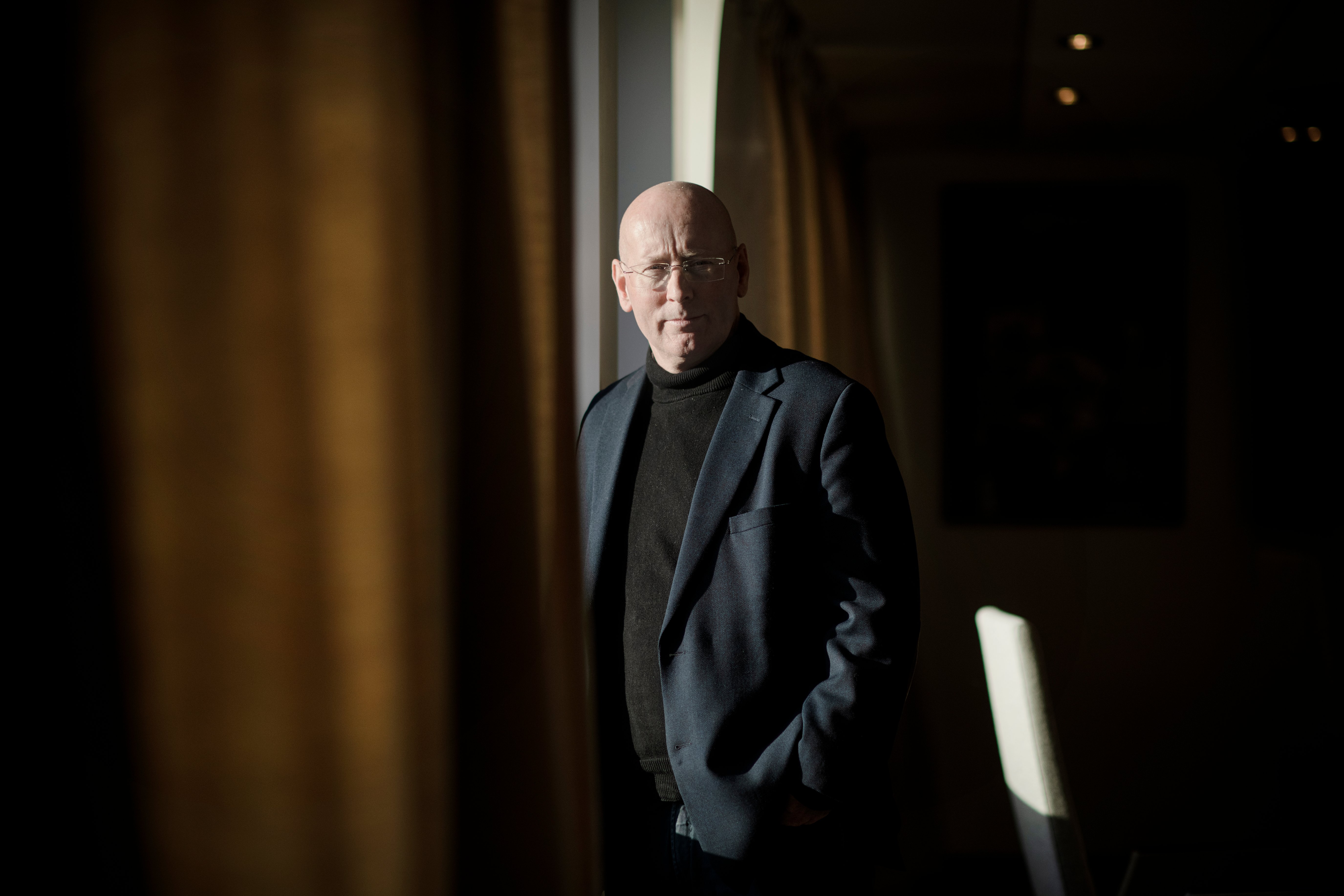 Mann i dress står ved vindu, mellom gardiner og ser mot kamera. Foto