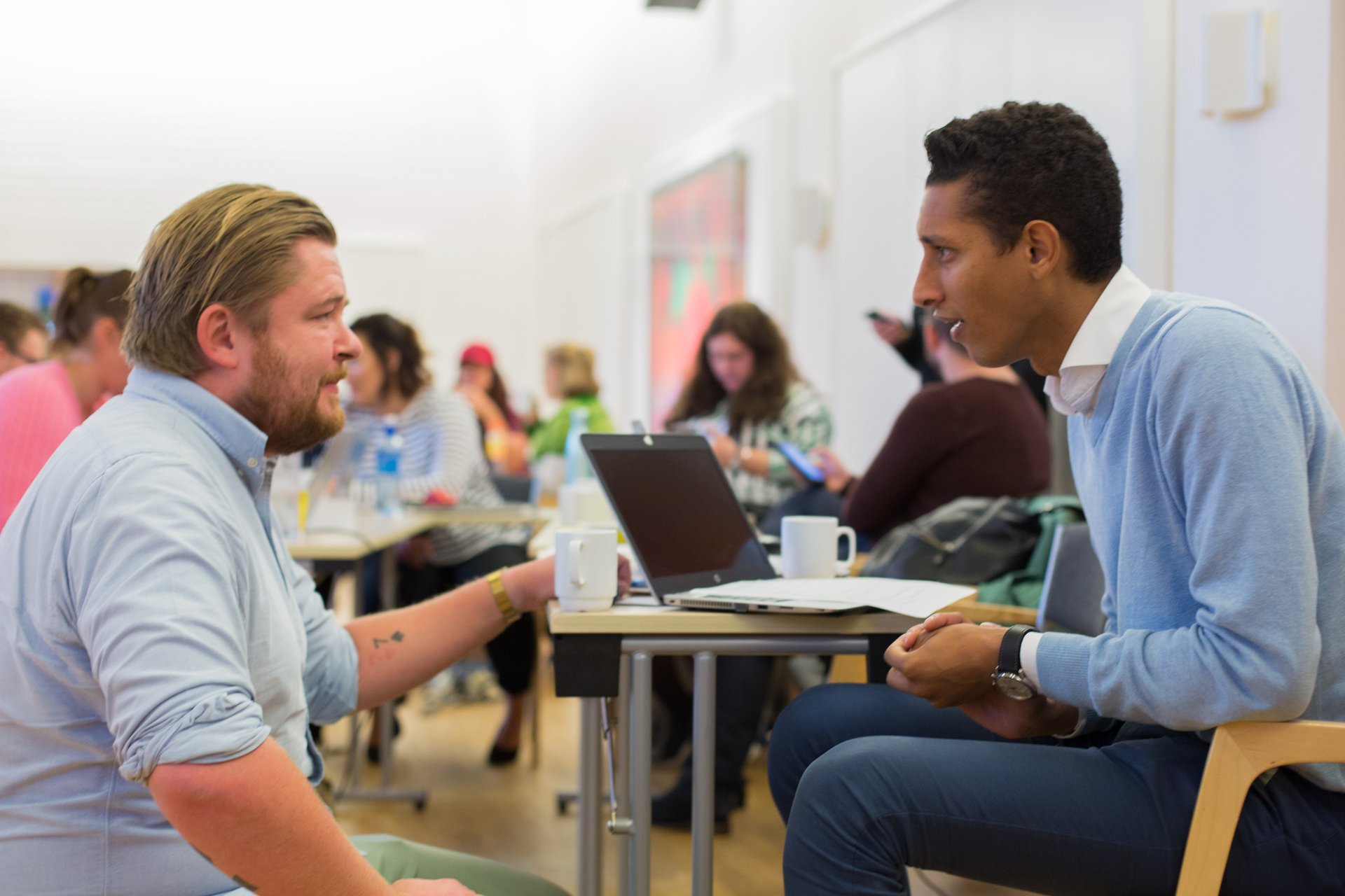To menn som sitter i et klasserom og snakker med hverandre ved en PC. Foto.