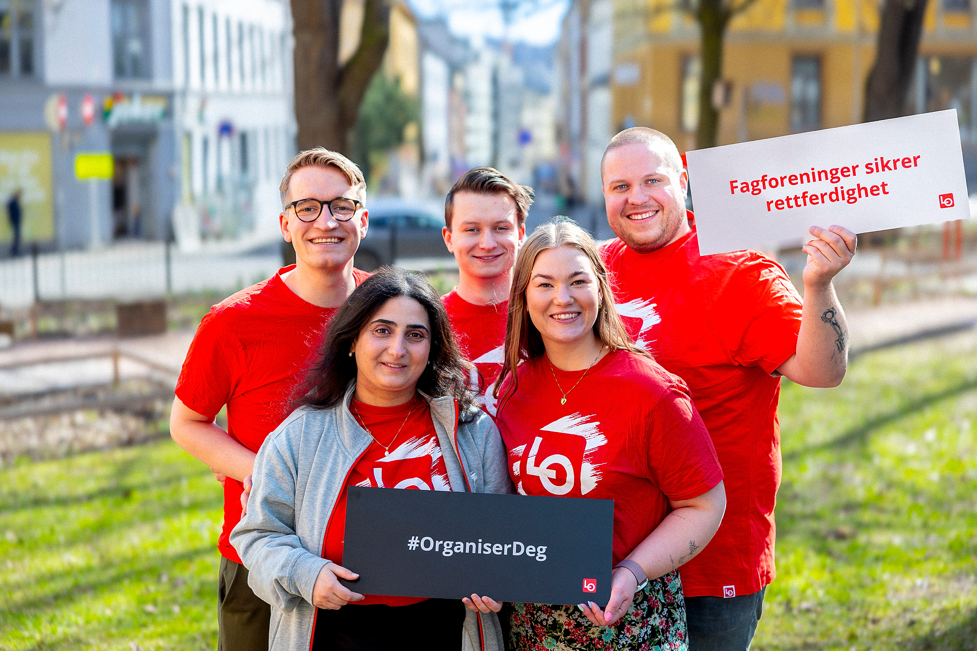 Fem ungdommer står samlet i matchende rød LO-tskjorte og holder opp plakater om fordeler ved å være fagorganisert. 