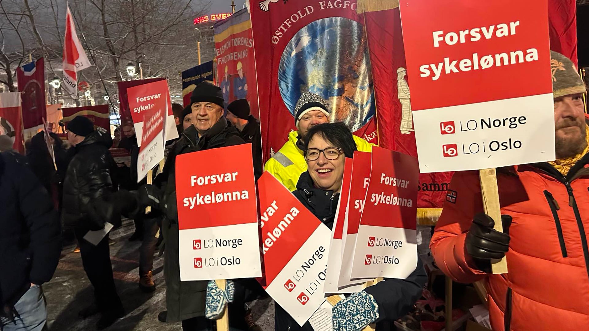 Mennesker foran faner som markerer mot kutt i sykelønna. Foto.