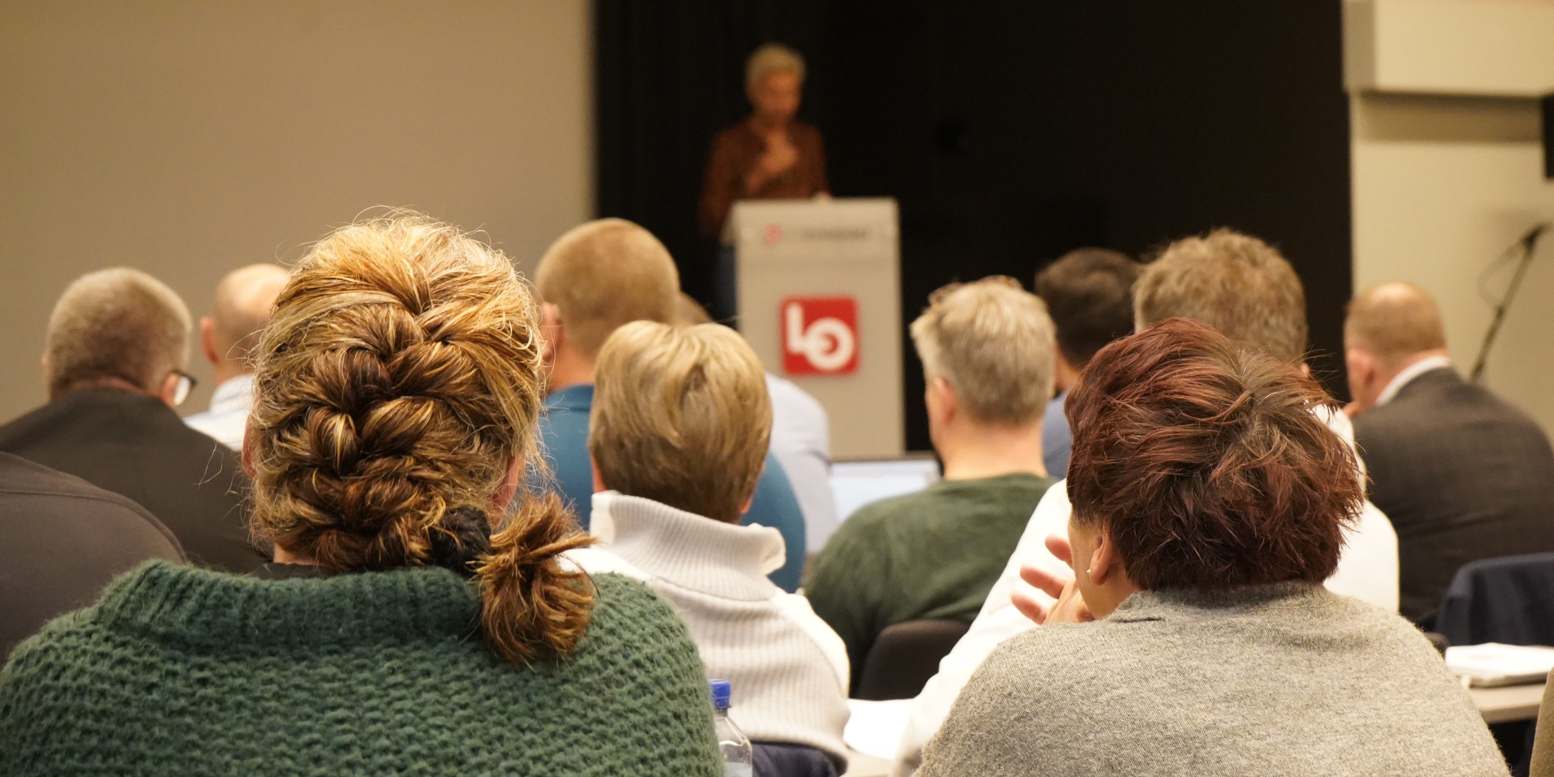 Møtebilde fra bakerst i salen, med lyttende forsamling under en innlegg på talestolen