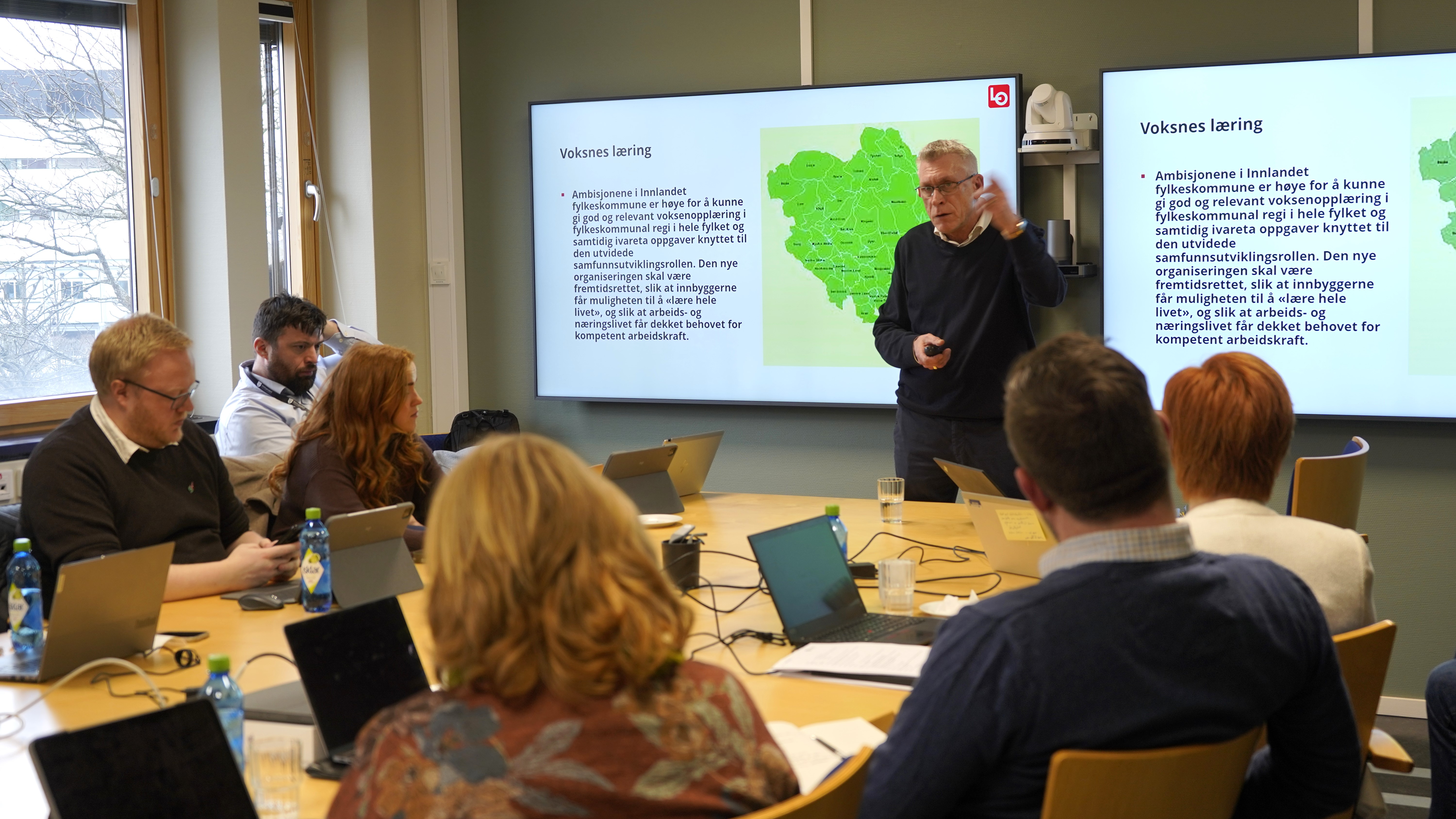 Iver Erling foran to framvisningsskjermer med utvalg for utdanning sittende rundt møtebordet