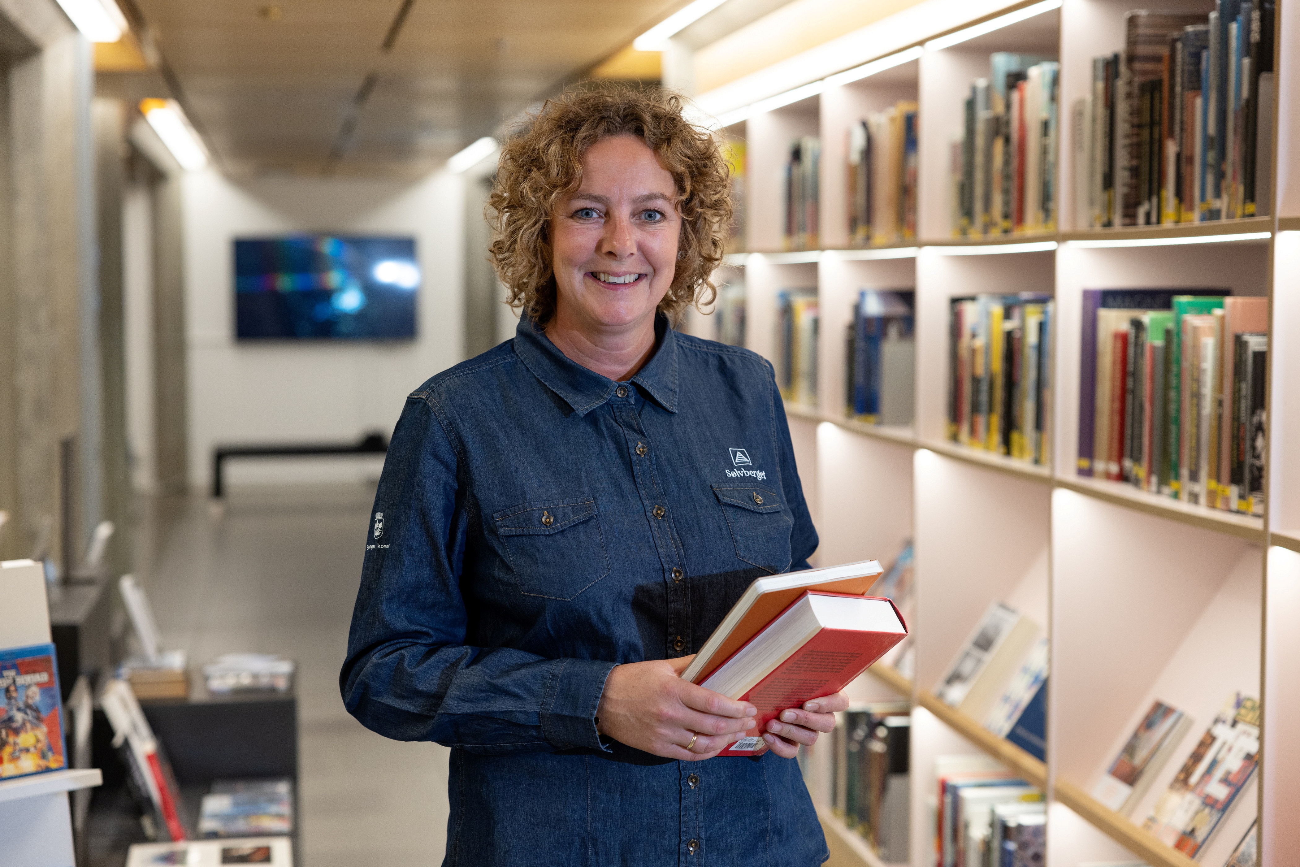 Bilde av en lærer står vendt med ansiktet mot kamera og med bøker i hendene. Bøker i hyllene bak. 