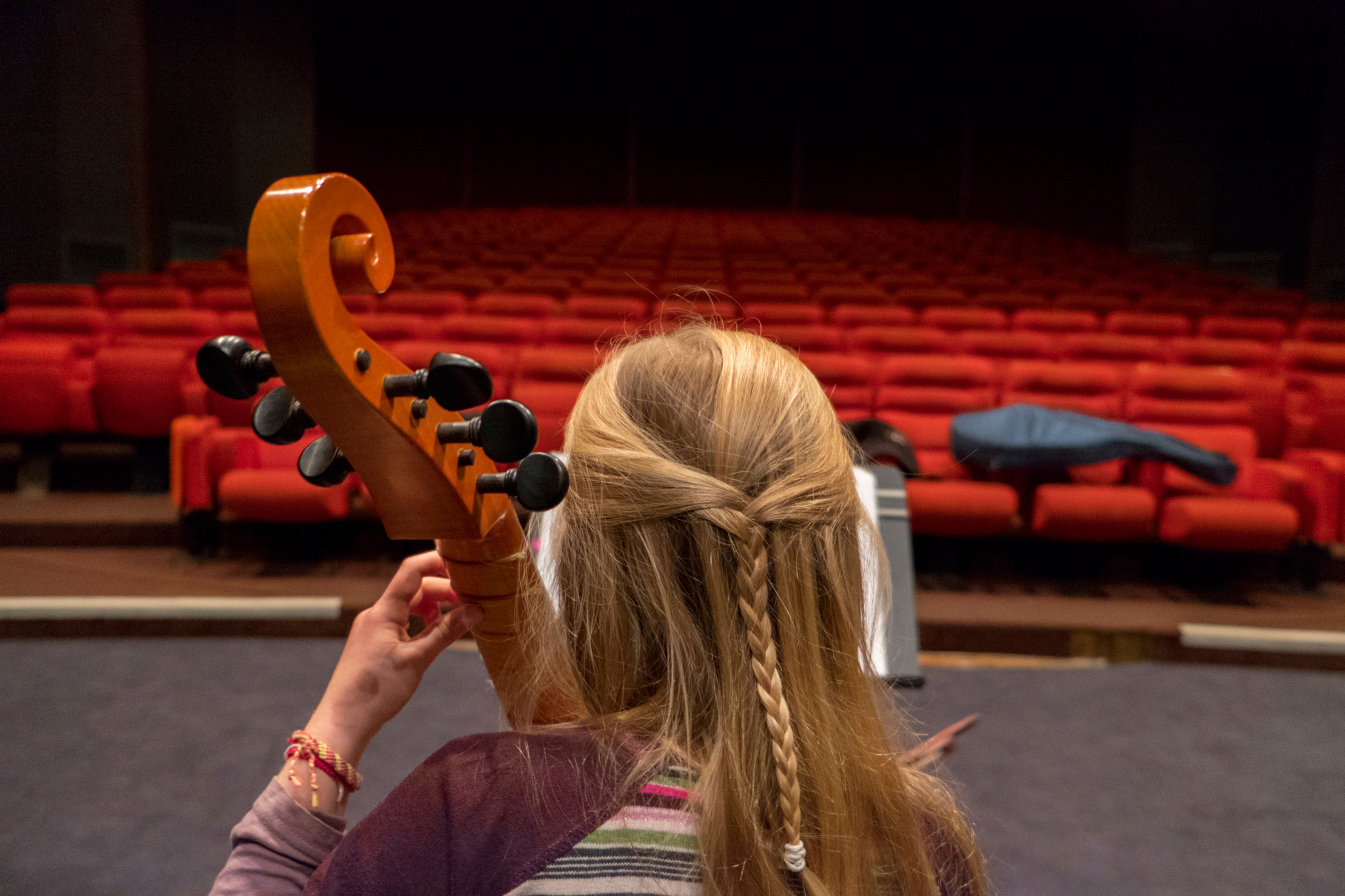 Kvinne som spiller strenginstrument sett bakfra foran en tom sal. Foto.