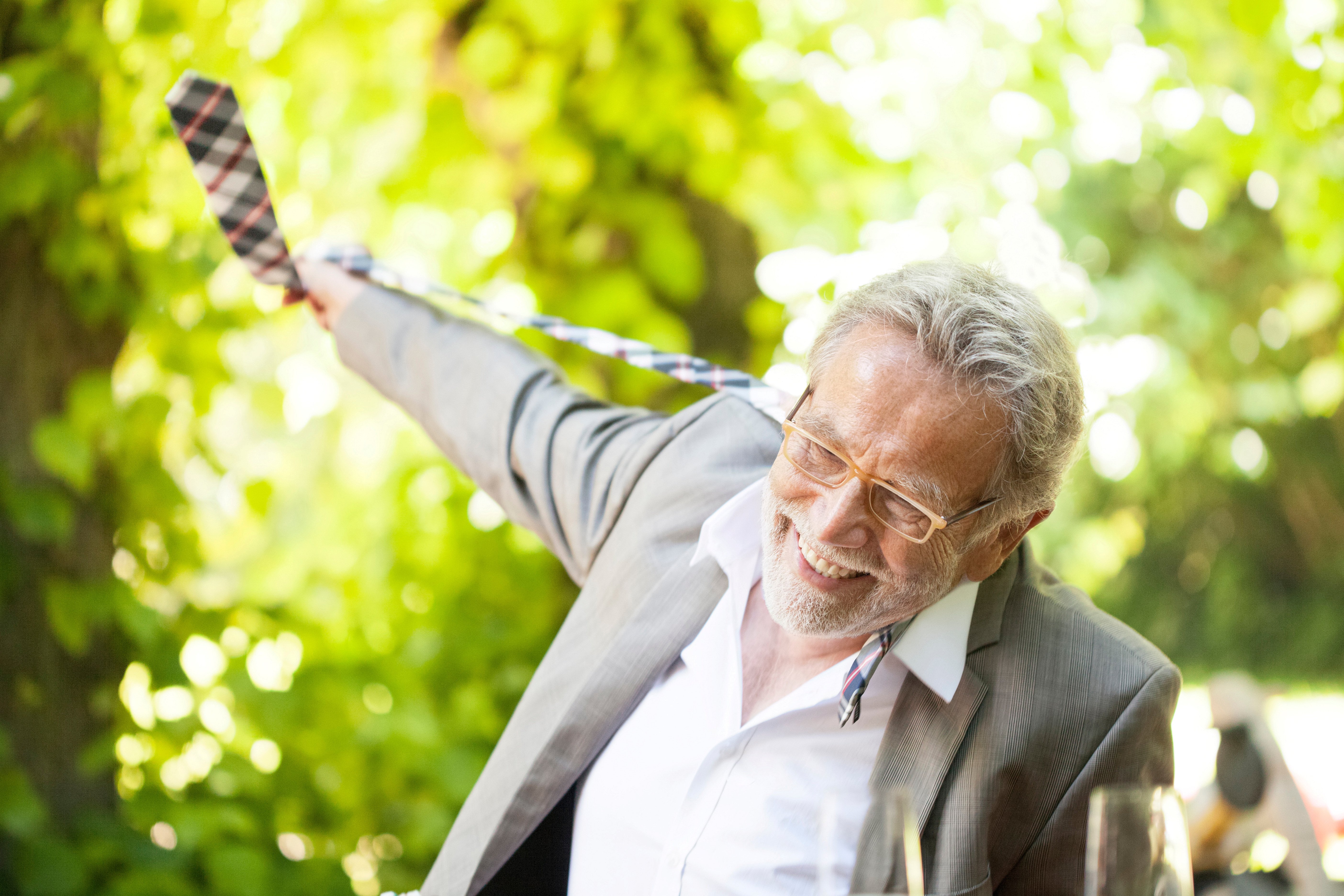 Eldre mann slår ut med armene og smiler ute i grønn natur. Foto.