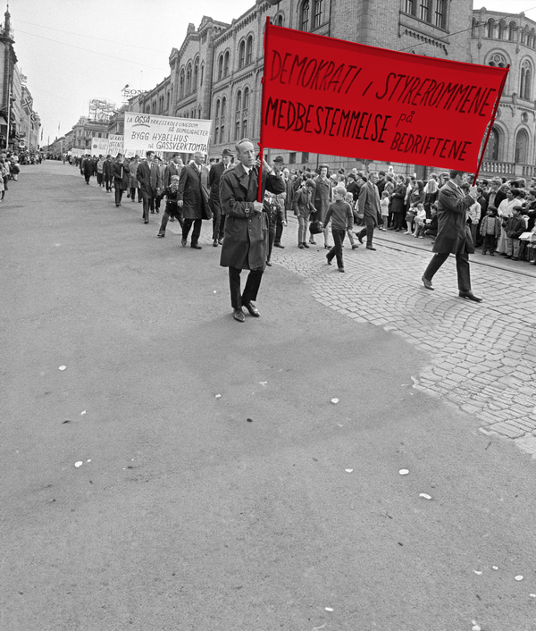 Bildet viser 1.mai-toget fra 1971 marsjere nedover Karl Johans gate. De to fremst i toget bærer en parole hvor det står "Demokrati, styrerommene, medbestemmelse på bedriftene".på 