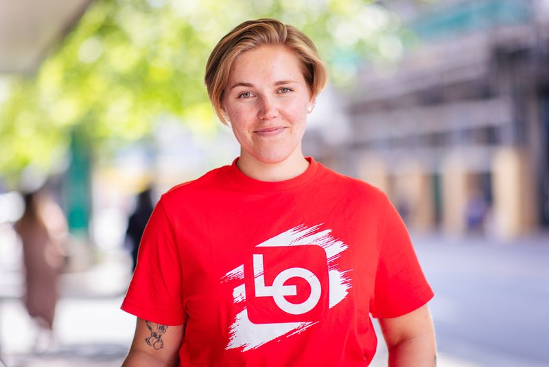 Bildet viser en ung dame ikledd rød LO t-skjorte. Med et smil om munnen og glimt i øyet, inviterer hun deg med til å bli med i kampen om framtiden.