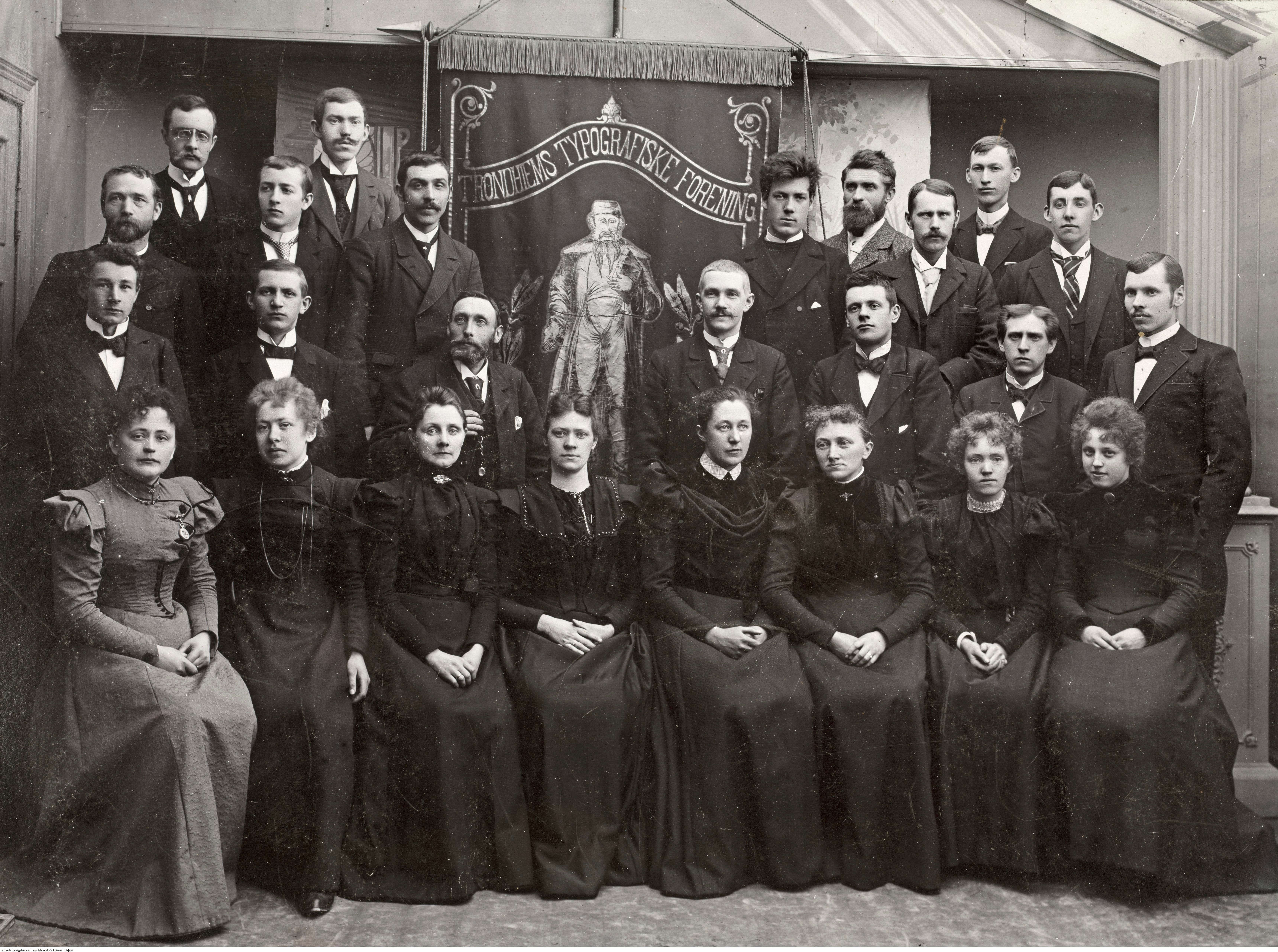 Bildet viser Trondheims typografiske forening, 1899. Menn og kvinner står samlet iført pene klær som sorte kåper og kjoler. 